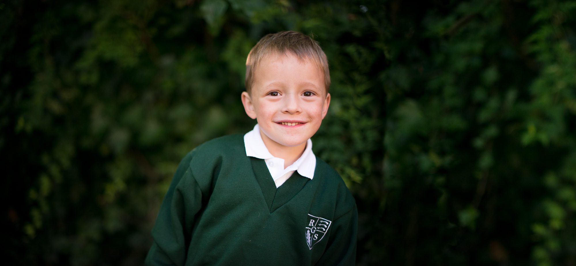 Outdoor Nursery school portrait of 4 year old boy in green uniform jumper in front of trees