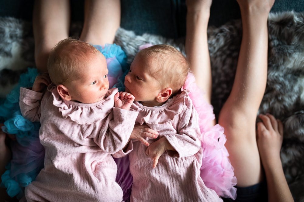 Twin newborn babies portrait in Pink dresses on their siblings knees at home